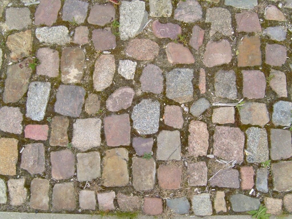 A cobblestone pavement seen from above: stones of several sizes and colours packed edge to edge in rough rows, with gaps and misfits
