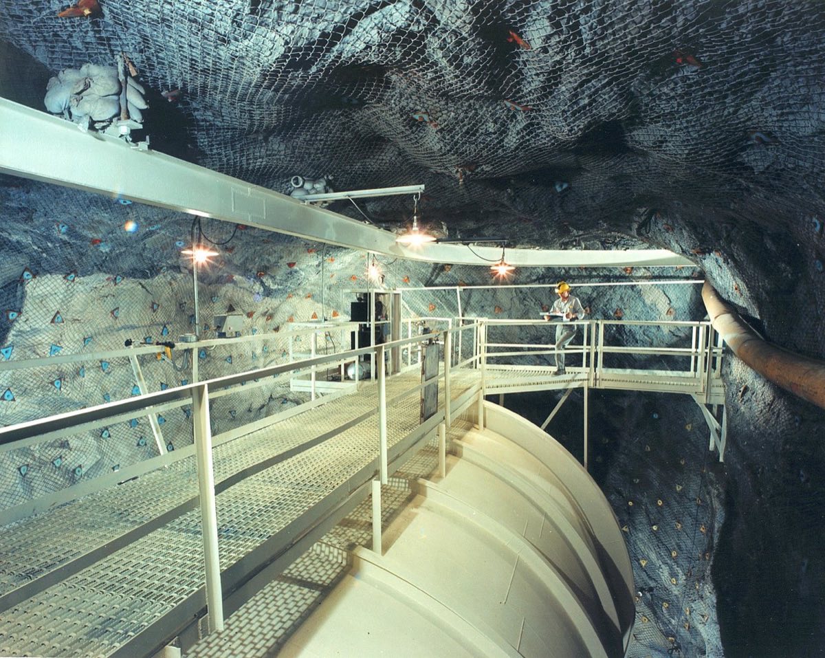 A curved pale steel tank with walkways over it, in a rock cavern lined with safety netting; a worker stands on the walkway