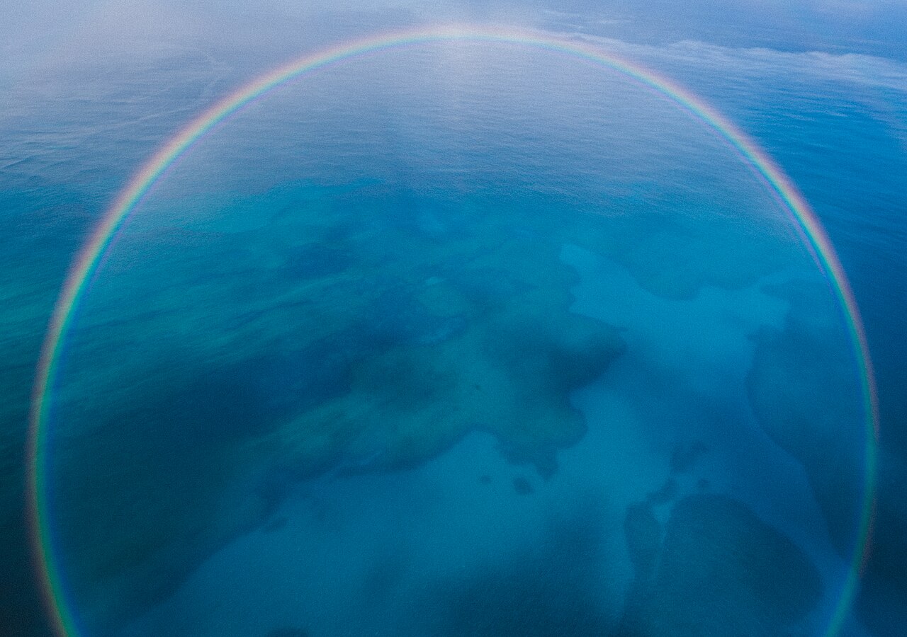 A nearly complete ring of rainbow seen from an aircraft above shallow sea, cut off only by the bottom of the frame